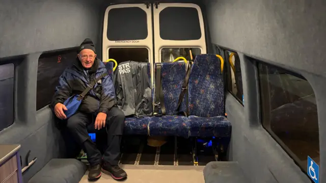 Un hombre con sombrero negro y chaqueta impermeable azul marino y negra. Está sentado en un autobús con asientos estampados en azul.