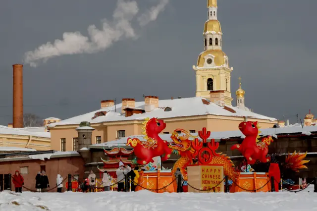 Инсталляция из двух красных лошадей и дракона между ними стоит возле стен крепости в снегу, чуть позади стоит миниатюрная китайская пагода. На заднем фоне видно православный храм. Возле китайской инсталляции прогуливаются люди. Все это огородено цепями, которые крепятся на столбах.