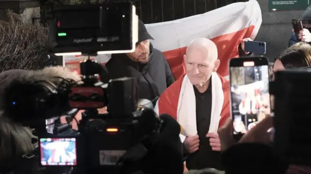 
紅白の旗を背に、紅白の旗を肩にかけたビャリャツキ氏が笑顔で中央に立ち、その周りを報道陣の複数のカメラが囲み、撮影している