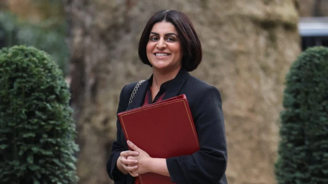 Home Secretary Shabana Mahmood dey waka outside for Downing Street. She wear one high-necked black jacket, black bobbed hair, and hold one burgundy folder.