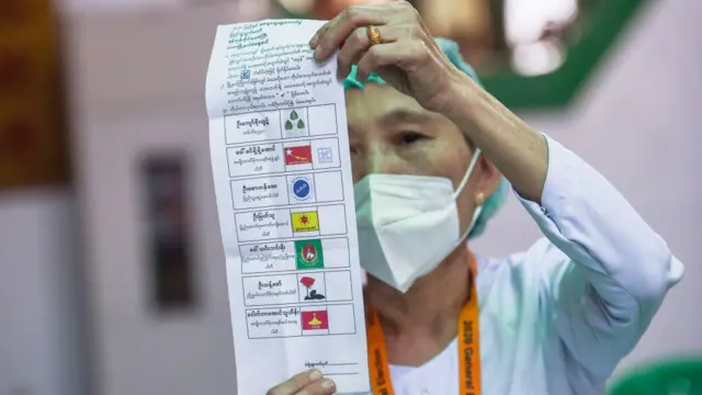 ပါတီရုပ်ပုံနဲ့ နာမည်တွေ ပါတဲ့ မဲစာရွက်ကို ထောင်ပြနေတဲ့ မက်စ်တပ်ထားတဲ့ အမျိုးသမီးတစ်ဦး 