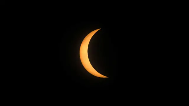 Foto colorida mostra eclipse solar, com o Sol na partefoguetinho da betanotrás, amarelo, e a lua o encobrindo quase inteiramente, como uma sombra