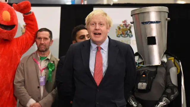 Boris Johnson aparece en el centro de la foto, con un traje negro, camisa azul claro y corbata roja de lunares. Detrás, a su derecha, está el Conde Cara de Bote de Basura.
