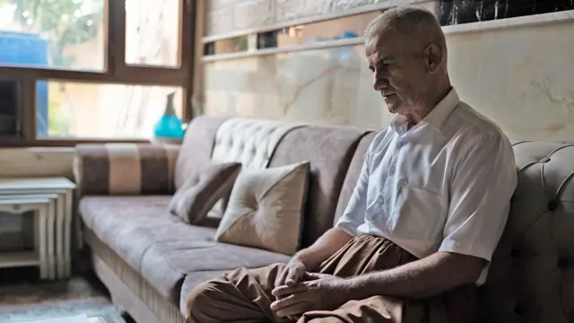 صورة لرجل يرتدي قميصاً أبيض، يجلس على أريكة بنية كبيرة، ويداه مطويتان في حجره، وهو يحدق إلى الأمام.