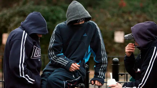 Trojica momaka pod kapuljačama sede i razgovaraju, ne vide im se lica, samo oči, trenerke su tamnoplave sa belim ili šarenim linijama na rukavima
