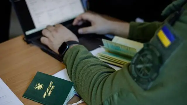 Військовий квиток лежить на столі, військовослужбовець заповнює персональні дані у нотубуку. 