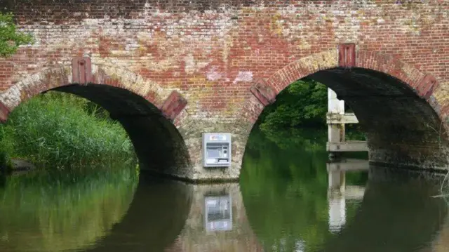 Dos arcos de ladrillo rojo se reflejan en el río con el atm gris unido a un contrafuerte central.