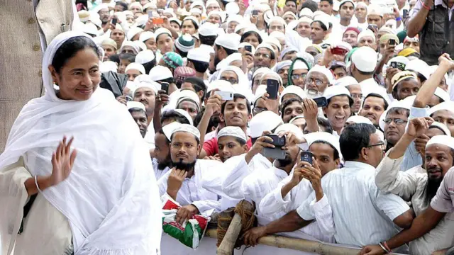 ममता बनर्जी अपना सिर ढके हुए एक कार्यक्रम में उनके पीठ के तरफ़ बहुत सारे मुस्लिम आदमी स्मार्टफ़ोन्स से उनकी तस्वीरें खींच रहे हैं