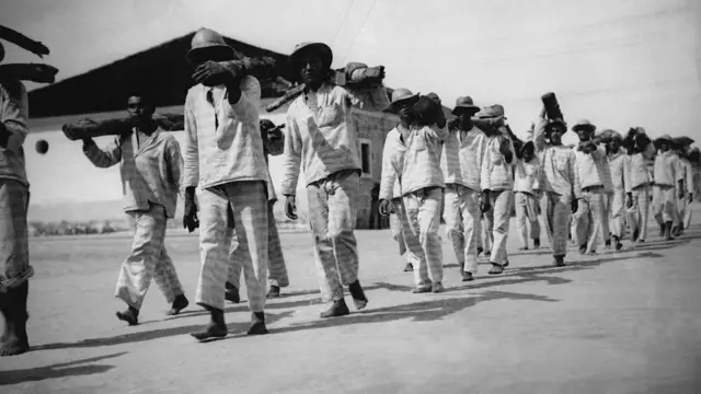 Homens negros com uniformes listrados carregam pedaços de madeira em fila 