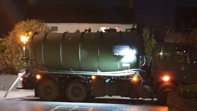 Un camión cisterna opera de noche bombeando aguas residuales de una alcantarilla en la carretera. Hay luces en el camión cisterna y una casa al fondo tiene una luz encendida en el piso superior.