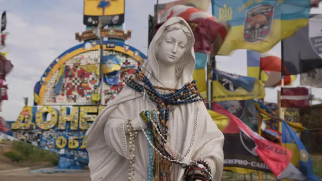 Une statue de femme est drapée de colliers de perles de rosaire, avec derrière elle des drapeaux de brigades d'unités ukrainiennes drapés sur des panneaux de signalisation.