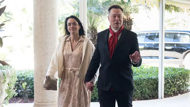 Shivon Zilis, Elon Musk ile el ele tutuşuyor. Zilis açık renk bir elbise ve ceket giymiş, Musk ise takım elbise ve kırmızı bir fular takmış. İkisi de gülümsüyor.
