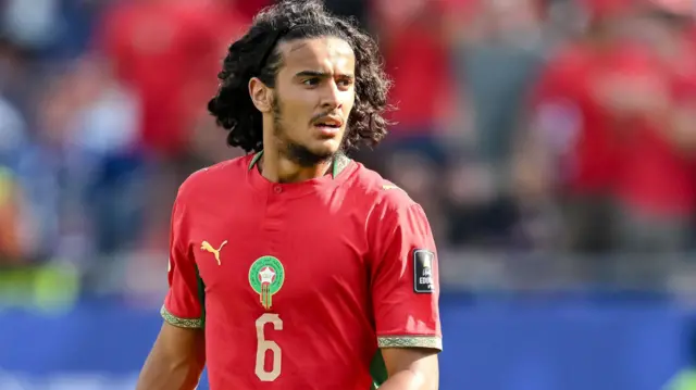 Ayoub Bouaddi, dont le buste est visible, vêtu d'un maillot rouge du Maroc floqué du numéro six doré et de l'écusson de la fédération, regarde vers la gauche pendant un match de football. On aperçoit au loin, de façon floue, une tribune accueillant des supporters.