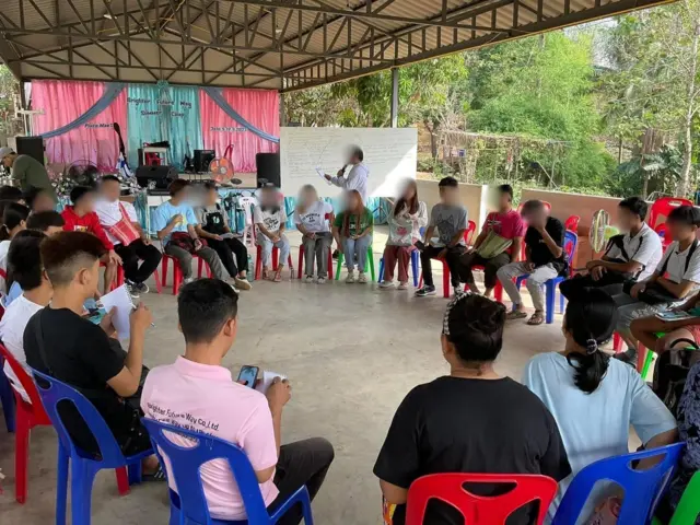 ၂၀၂၃ ခုနှစ်၊ မဲဆောက်မြို့မှာ Brighter Future Way ကုမ္ပဏီက လူငယ်တွေကိုစခန်းသွင်းလေ့ကျင့်ခဲ့တဲ့ မြင်ကွင်း၊ အမိုးတစ်ခုအောက် ကွန်ကရစ်ကြမ်းပြင်ထက် ပလပ်စတစ်ထိုင်ခုံတွေပေါ်မှာ လူငယ်အမျိုးသား အမျိုးသမီး ၂၀ နီးပါး ဝိုင်းပတ်ထိုင်နေပါတယ်။ သူတို့ အနောက်ဘက်မှာ စင်မြင့်တစ်ခုကို မြင်တွေ့ရပါတယ်။ အဆောက်အဉီ အမိုးအနောက်ဘက်မှာတော့ သစ်ရိပ်ဝါးရိပ်ကို မြင်တွေ့ရ၊ ပလပ်စတစ်ထိုင်ခုံတွေတွကတော့ အနီ၊ အပြာ အများစုဖြစ်တာကို မြင်တွေ့ရတယ်။ ကင်မရာရှေ့က လူ ၆ ဦးထက်မနည်းကတော့ ကင်မရာကို ကျောပေးနေ 