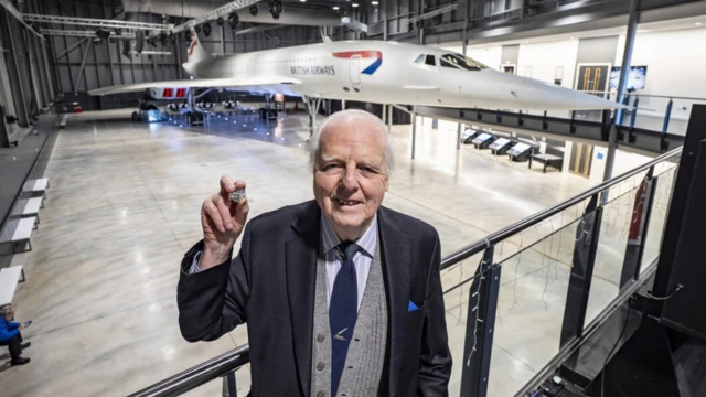 John Britton, Concorde's chief engineer, holds the new coin in his uplifted hand, standing on a platform in the hangar with Concorde behind him. He is smartly dressed in a blue jacket, striped shirt and tie. 