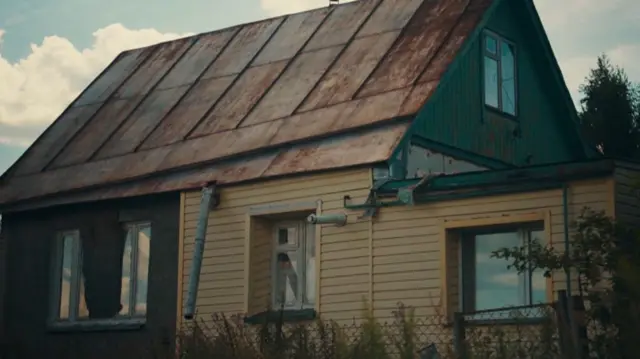 Uma casa de madeira nas cores verde e amarela, ladeada por plantas. O vidro de uma das janelas está quebrado, e a cobertura parece enferrujada.