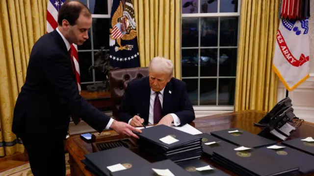Trump signe des mesures exécutives au bureau de la Résolue, dans le bureau ovale. Des piles de documents sont posées sur le bureau, chacun portant un Post-It indiquant de quoi il s'agit. Un assistant soulève un document tandis que Trump utilise un stylo noir épais pour signer son nom sur un autre document.