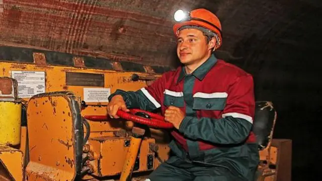 Рабочий в красно-бело-серой спецовке и оранжевой каске с фонариком. Он за рулем специального грузового транспортного средства - оно тоже оранжевого цвета - в шахте по добыче калия
