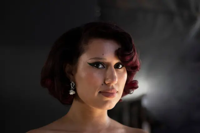 A portrait of Raye looking into the camera, her face slightly turned to her left. She wears heavy thick eyeliner and curled red hair.
