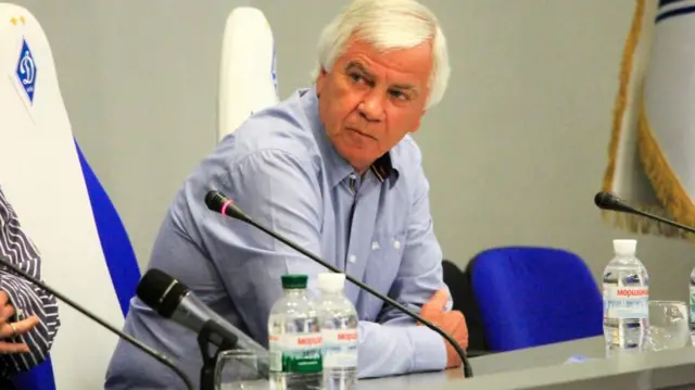 Володимир Мунтян у блакитній сорочці сидить за столом, дивиться убік, на столі стоять пляшки з водою