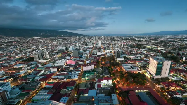 Una foto aérea de San José, Costa Rica. Parace un final del atardecer, se ven ya algunas luces encendidas en parques y avenidas, con una montaña de fondo.