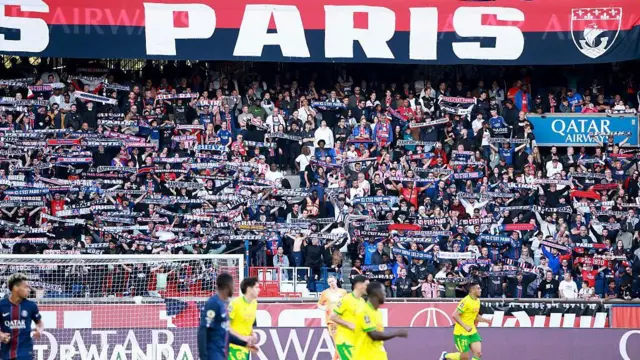 لاعبو باريس سان جيرمان (اللون الأزرق) ولاعبو (نانت) وخلفهم قسم من مدرج جمهور باريس يرفع لافتة ضخمة بعبارة باريس باللغة الفرنسية.