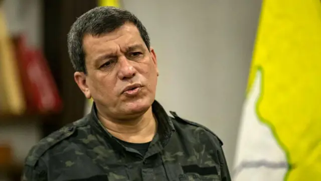 Mazlum Abdi, üzerinde bir askeri kıyafetle açıklama yapıyor. 