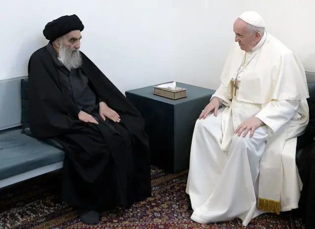 solda baştan aşağı siyah kıyafetiyle sistani, sağda baştan aşağı beyaz kıyafetiyle papa, birbirlerine bakarak ve oturarak sohbet ediyor