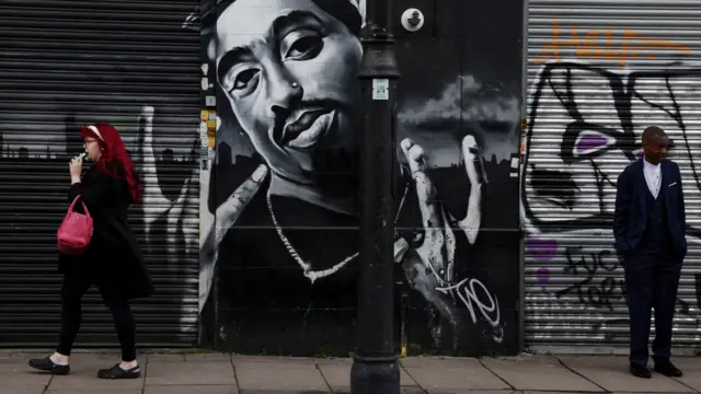 Uma mulher caminha enquanto outra pessoa está diante de um mural do astro do hip-hop Tupac Shakur, em Londres, em 2024