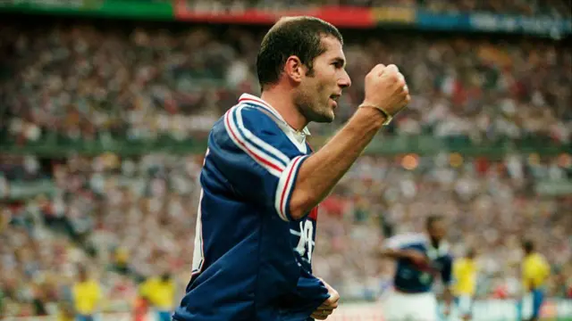 A imagem mostra um jogador da seleção francesa comemorando de forma vibrante durante uma partida, com o punho cerrado e expressão de entusiasmo. Ao fundo, um estádio lotado aparece desfocado, enquanto jogadores brasileiros podem ser vistos à distância, reforçando o contexto de um confronto internacional.
