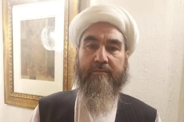 تصویر شیخ محمد قاسم راسخ با ریش بلند گندمگون و عمامه سفید، پیش روی یک تابلویی هنری ایستاده است