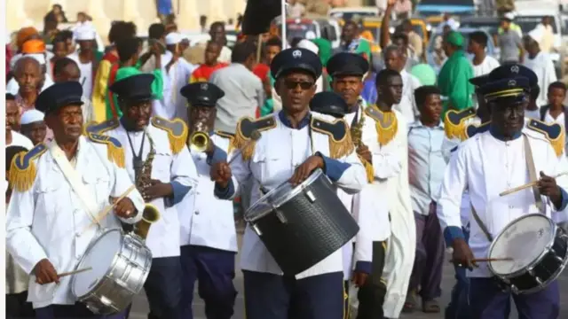 Aworan yii ṣafihan awọn ọkunrin to gbe ilu kọ ọrun ti wọn si n lu u, pẹlu awọn to n fun fere. Oju popo ni wọn wa, ero pọ lẹyin wọn, aṣọ funfun ati ṣokoto dudu ni awọn onilu ati onifere naa wọ.