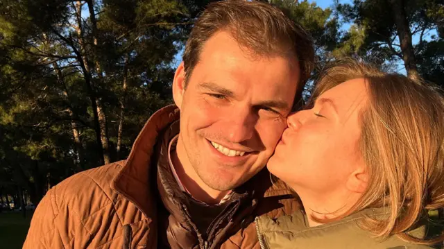 Dwie osoby stoją blisko siebie na pierwszym planie, ubrane w pikowane kurtki, jedną brązową i jedną zieloną. Znajdują się na zewnątrz, w zalesionym terenie, za nimi rosną wysokie sosny. Ciepłe światło słoneczne pada na scenę, a w tle widać błękitne niebo prześwitujące przez drzewa.