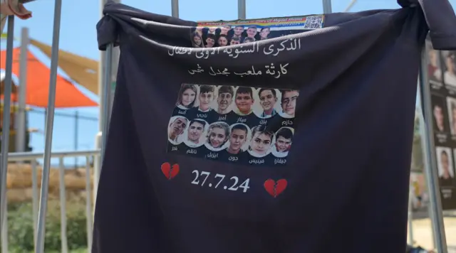 قطعة من قماش أسود مطبوع عليها صور وأسماء 12 طفلاً وقد علقت على سور معدني. وقد طبع أسفل قطعة القماش صورتان باللون الأحمر لقلبين مكسورين وتاريخ 27.7.24 باللون الأبيض.