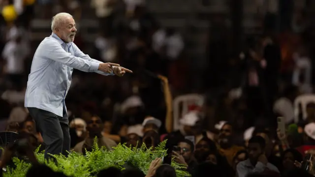 O presidente Lula em um palco apontando os indicadores das duas mãos para o público.