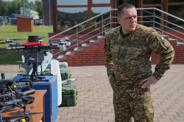 Александр Пивненко стоит в военной форме на улице возле нескольких беспилотников