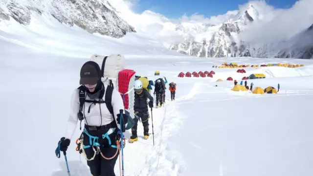 Integrantes de uma equipe de expedição caminham com dificuldade pela neve entre o Acampamento 1 e o Acampamento 2 durante uma subida de aclimatação. Ao fundo, há dezenas de barracas montadas