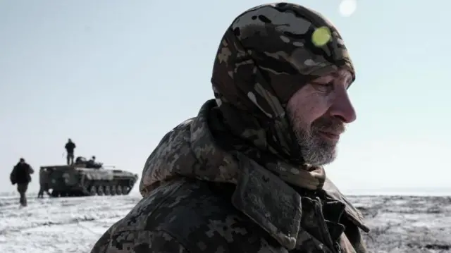 من القوات الأوكرانية يرتدي زياً عسكرياً وجاكيت شتوي بنفس اللون الزيتي العسكري وخلفه مركبة عسكرية واثنين من زملائه. في مكان يبدو مليئاً بالثلج.