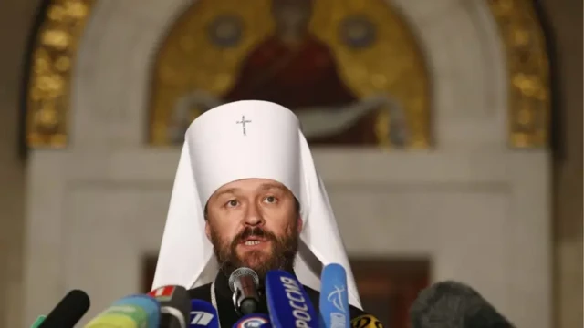 Іларіон в чорному релігійному одязі з білим головним убором стоїть перед мікрофонами і виступає на пресконференції або брифінгу. На задньому плані — за інтер’єр церкви з теплими кольорами і розмитим зображенням ікони.