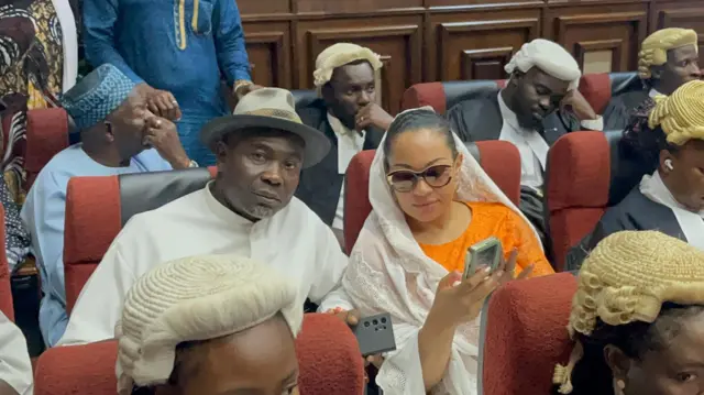 Senator Natasha husband looking at di camera while di wife dey press her phone, some lawyers dey show for di background