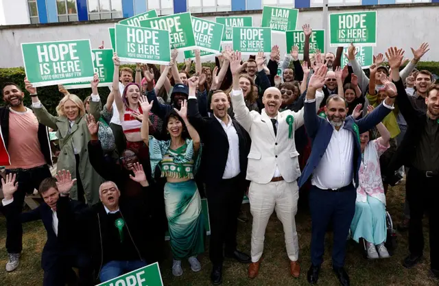 Grupa uśmiechniętych osób trzymających zielone transparenty z napisem 'Hope is here' (ang. Nadeszła nadzieja) stoi na trawie przed betonowym budynkiem. Kilka z nich ma przypięte na piersiach zielone rozety z logo brytyjskiej Partii Zielonych. 