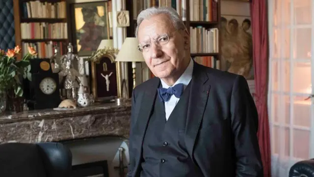 Michel Maffesoli, um homem branco idoso de terno, gravata borboleta e óculos em frente a prateleiras de livros 