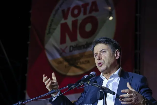 İtalyan politikacı Conte, beyaz bir gömlek ve lacivert bir ceket giymiş, iki eli açık bir şekilde önündeki mikrofona konuşuyor. Arkasında büyük bir poster var. Posterin üzerinde Hayır yazıyor. 