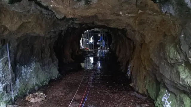 El interior de una cueva rocosa con tres cuerdas que discurren por el suelo húmedo. A lo lejos, dos rescatistas de montaña se ven iluminados tras una puerta metálica.