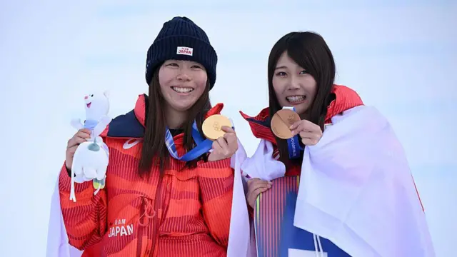 Золотая медалистка Мари Фукада и бронзовая медалистка Кокомо Мурасе из сборной Японии позируют для фото с флагом Японии во время церемонии награждения по сноуборду среди женщин в дисциплине слоупстайл на двенадцатый день зимних Олимпийских игр Milano Cortina 2026 в сноупарке Ливиньо 18 февраля 2026 года в Ливиньо, Италия.