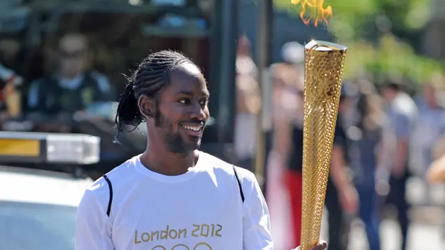 LONDON2012と書かれた白いTシャツを着たアーデイ氏が、金色の聖火トーチを持って道路を走っている