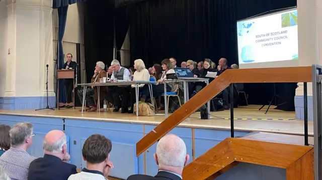 Un hombre en un pedestal está hablando mientras muchos otros se sientan a su lado en un escenario.