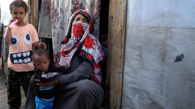 سيدة ترتدي نقاباً تجلس على الأرض وتمسك بطفل صغير تقف إلى جانبهما طفلة أكبر سناً