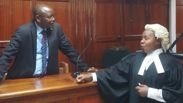 Umubikira Immaculate (iburyo) yambaye ikanzu y'umukara n'ingofero y'umweri arimo kubaza ibibazo umugabo wambaye isuti y'umukara n'itayi uri imbere ye