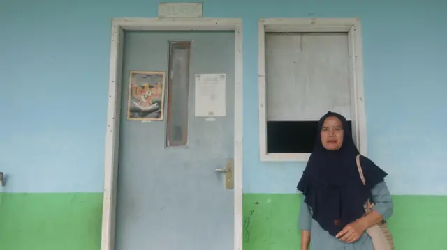 Sarkiyah memakai baju hijau dan jilbab sedada warna hitam, berdiri di depan tembok dengan pintu tertutup dan jendela.